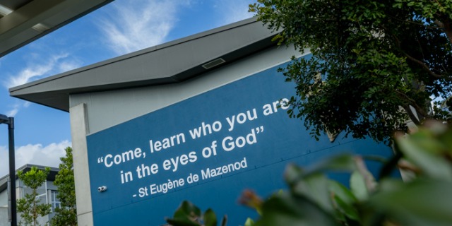 Exterior view of a St Eugene College building with a blue wall displaying the quote, “Come, learn who you are in the eyes of God – St Eugene de Mazenod.” The building is partially framed by trees and greenery under a blue sky, reflecting the college’s faith-based identity.