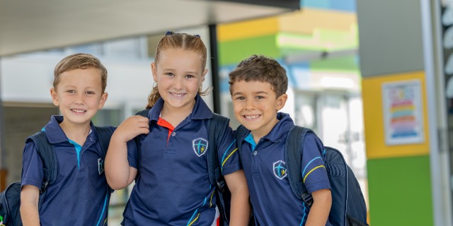Three smiling young students wearing St Eugene College uniforms stand together with their school bags, looking at the camera. The children are in an outdoor learning space on campus, conveying a welcoming and supportive early years school environment.