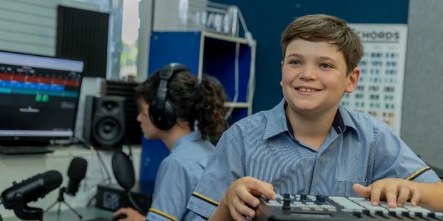 Middle years students work with audio and media equipment in a classroom learning space. One student smiles while operating a mixing controller, while another wears headphones at a computer workstation, representing creativity, collaboration and hands‑on digital learning at St Eugene College.