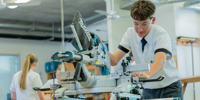 A senior student wearing safety glasses uses a power saw in a technology workshop at St Eugene College. Other students work in the background, highlighting hands‑on learning, practical skills and safe use of tools in the senior years.