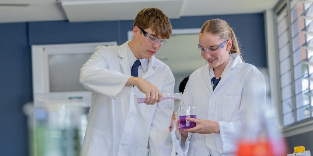 Two senior students wearing lab coats and safety glasses conduct a science experiment together in a laboratory. One student carefully pours liquid into a measuring cylinder while the other observes, representing collaborative learning and scientific inquiry in the senior years.