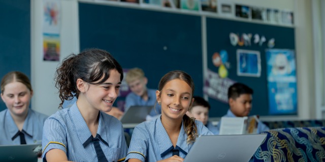Students work on laptops in a classroom learning space at St Eugene College. Two students sit together smiling while using a laptop, with other students working independently in the background, representing collaborative learning and the integration of digital technologies in everyday classroom practice.